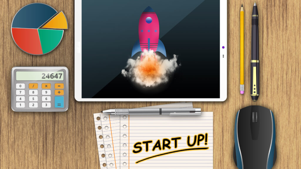 Start up represented as an iPad with a rocket ship on it along with a pen and pencil next to it. The words start up on a piece of paper with a pie chart and calculator.